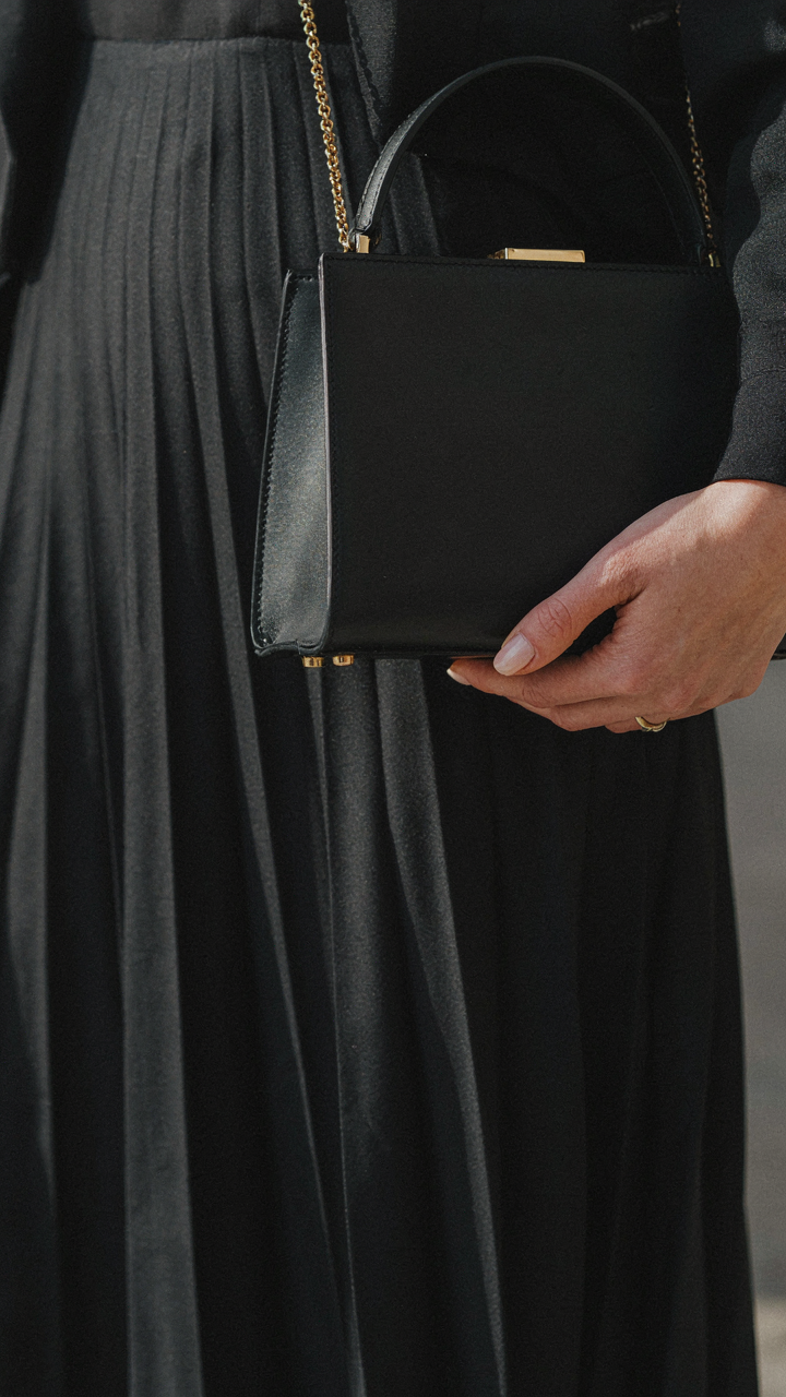 Woman holding a sleek, structured handbag against a dark pleated midi dress.