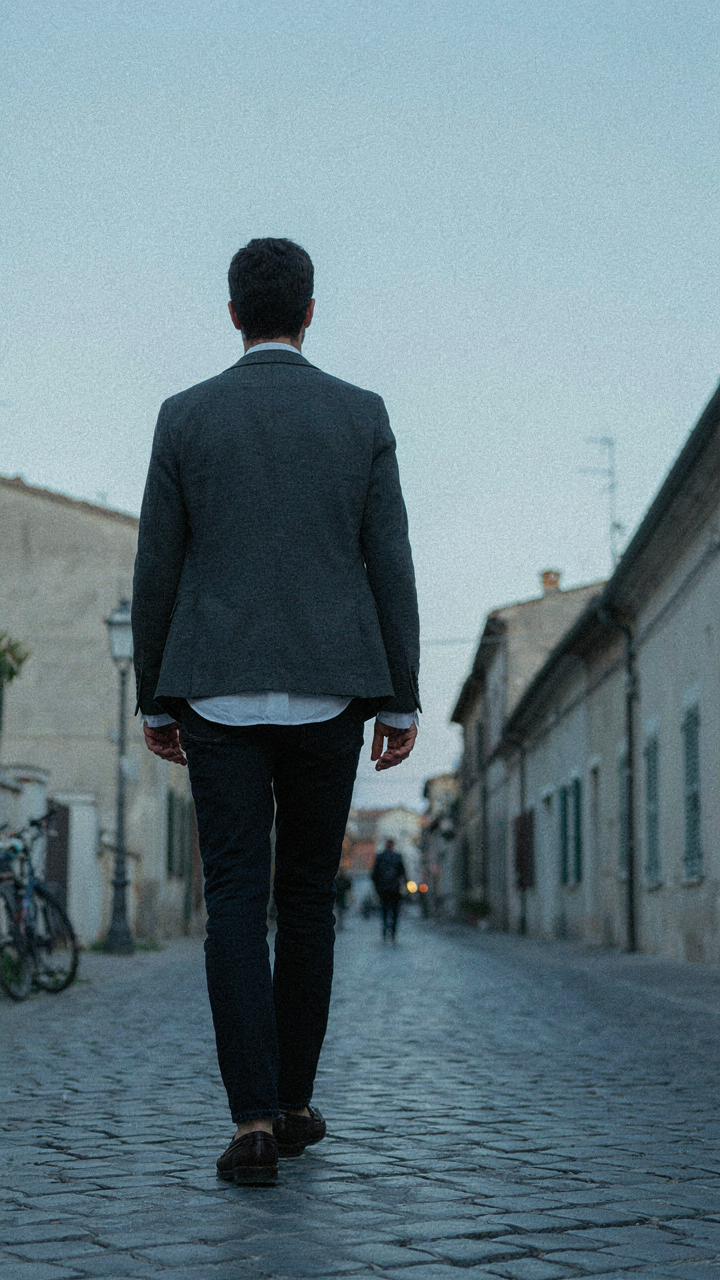 A person in dark jeans, a structured shirt, and a jacket walks away.