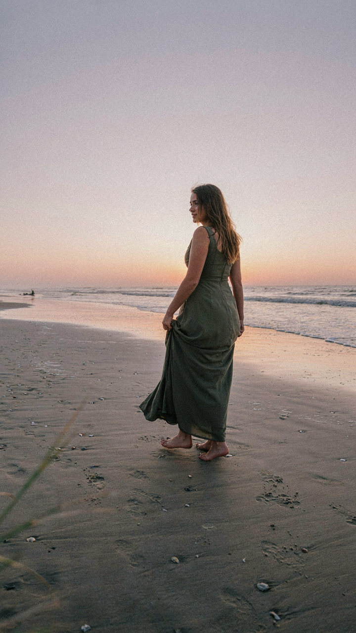A woman in an olive green maxi wedding guest dress on a beach at sunset.