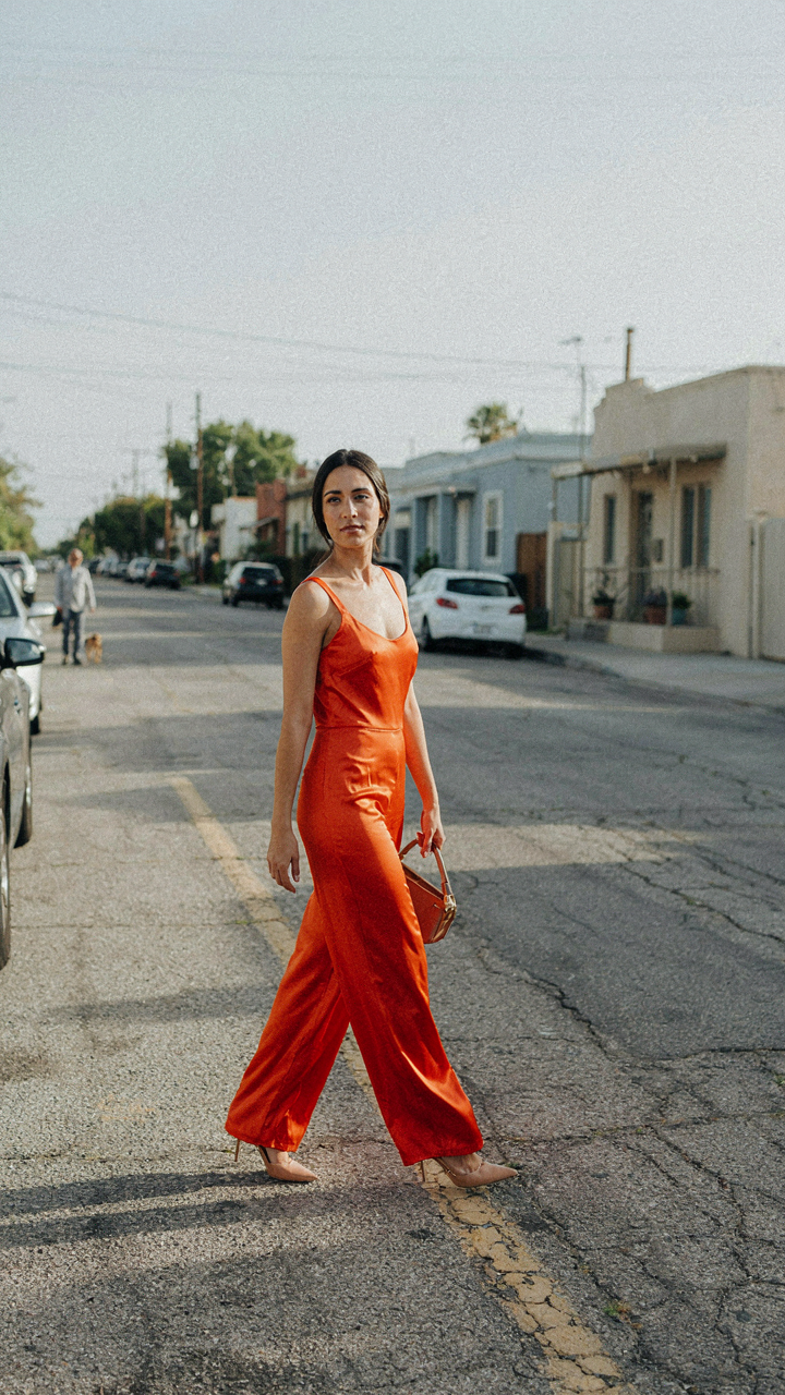 A woman in a vibrant orange satin jumpsuit walks confidently down a street.