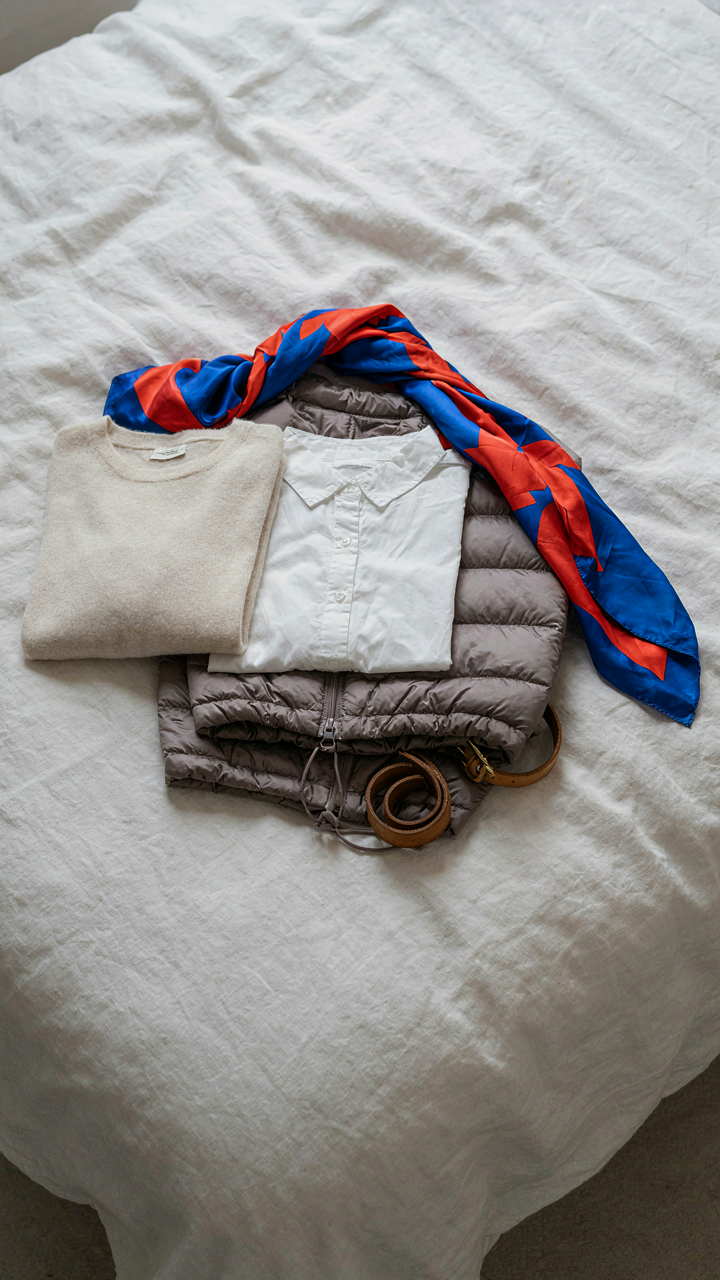 A women's travel capsule wardrobe on a bed, including a colorful silk scarf.