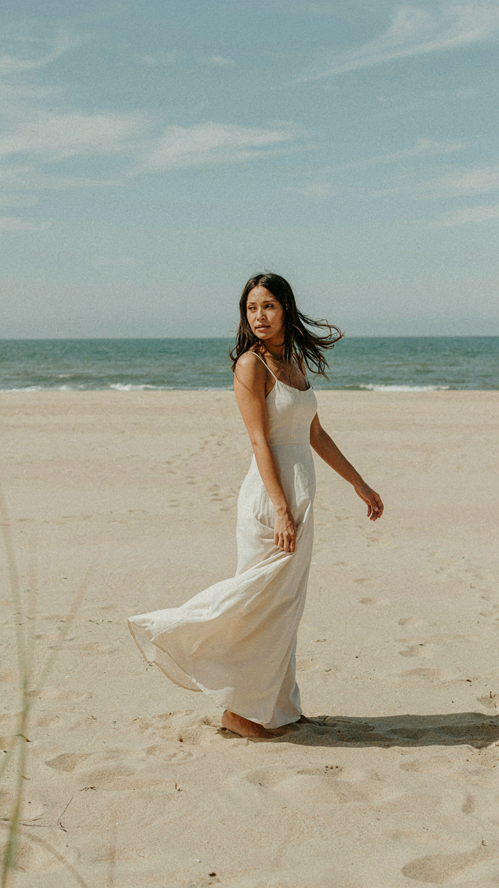 A woman in a flowing dress stands on a sandy beach looking at the ocean.