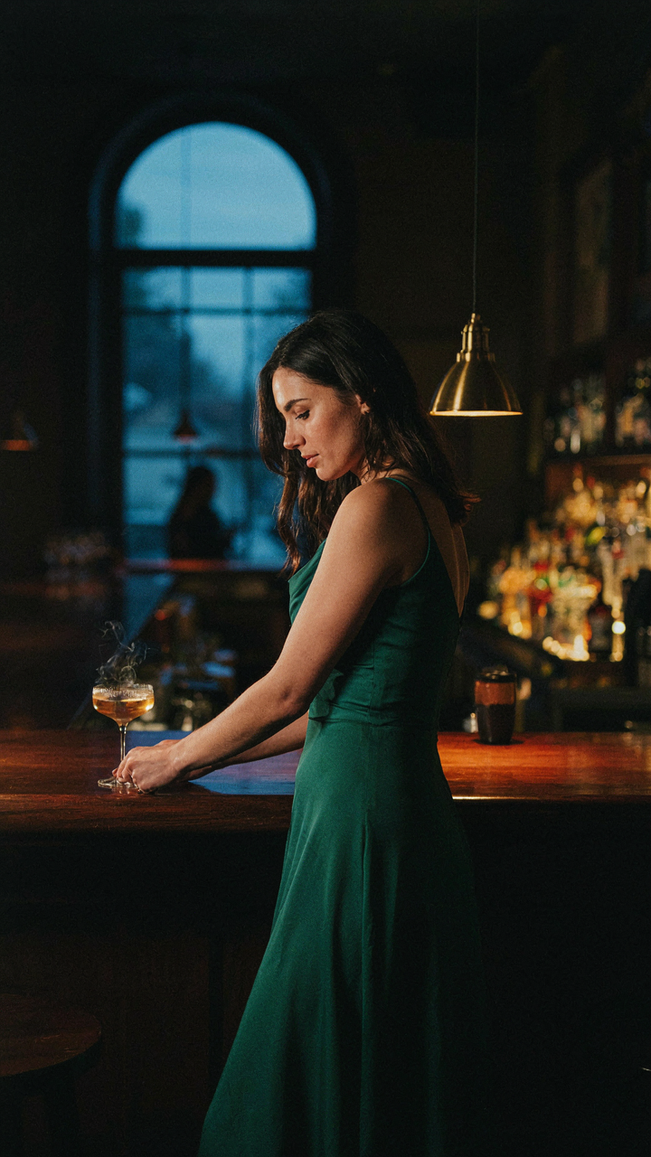 Woman in a flowing cocktail dress moves naturally, reaching for a drink.