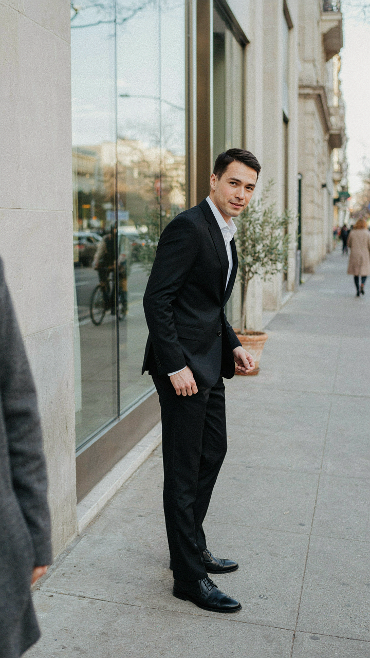 A person in a black suit leans casually, looking effortless and belonging.