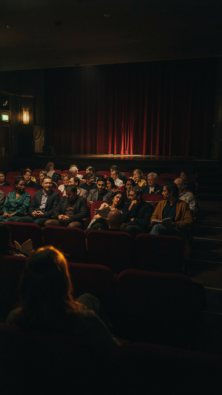 A theater audience watches a performance in a dimly lit room.