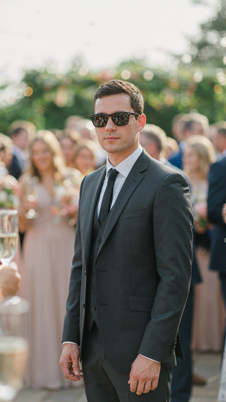 A wedding guest in formal attire wears dark, reflective sunglasses, visually detached from the
