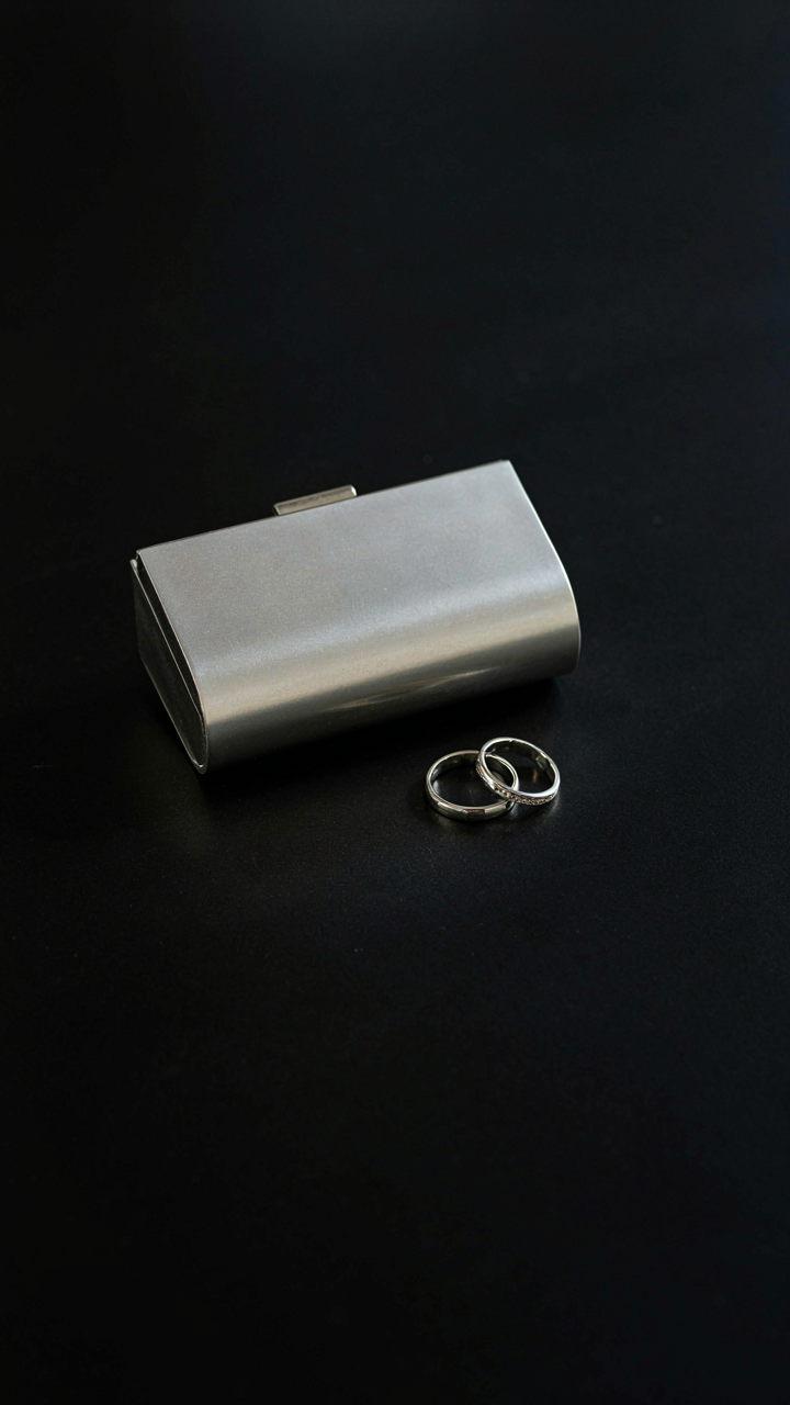 Minimalist silver clutch and two thin silver rings resting on a dark surface.