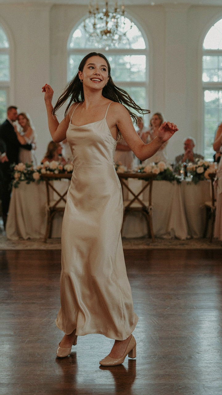 A woman in a satin dress and block heels dances joyfully at a reception.