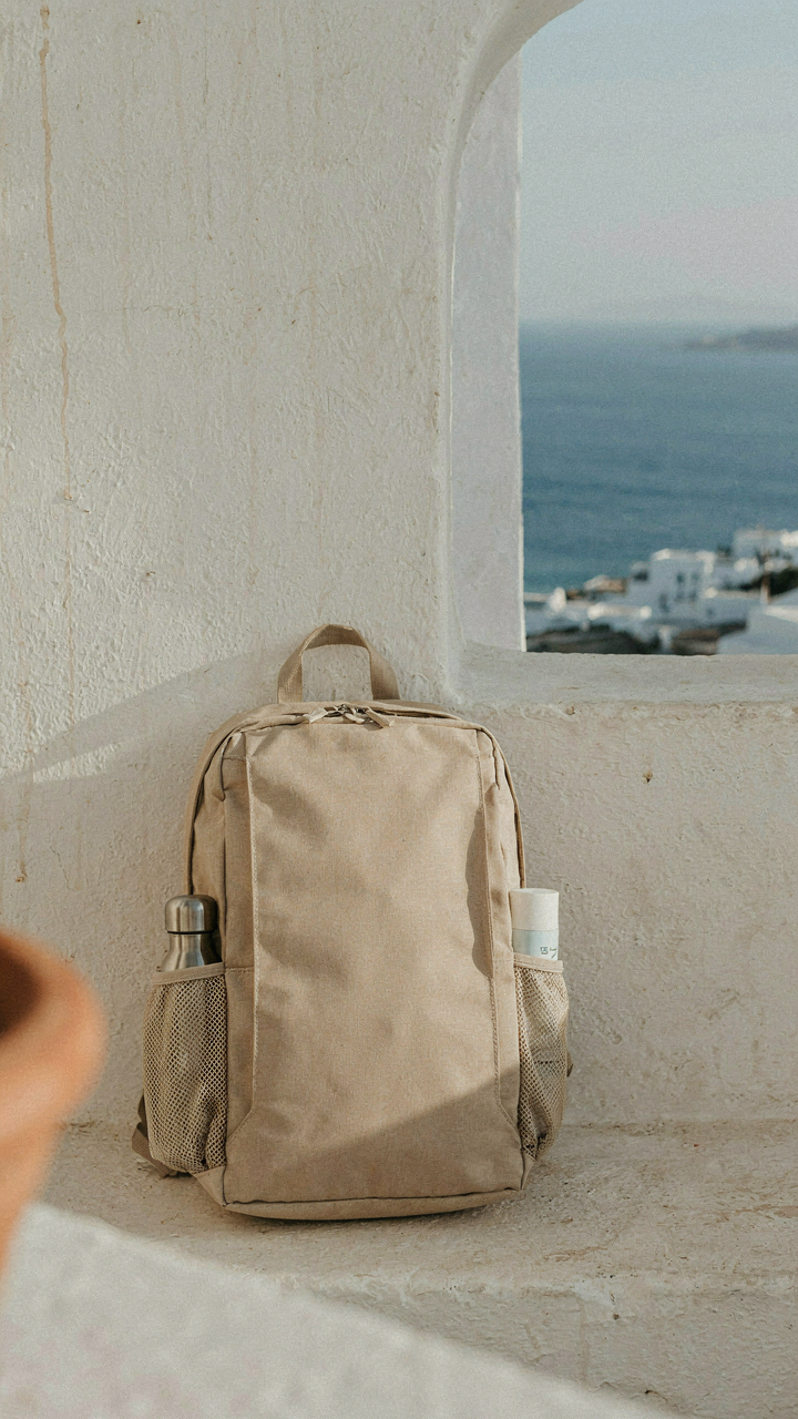 A travel backpack with a water bottle and sunscreen against a Greek island backdrop.