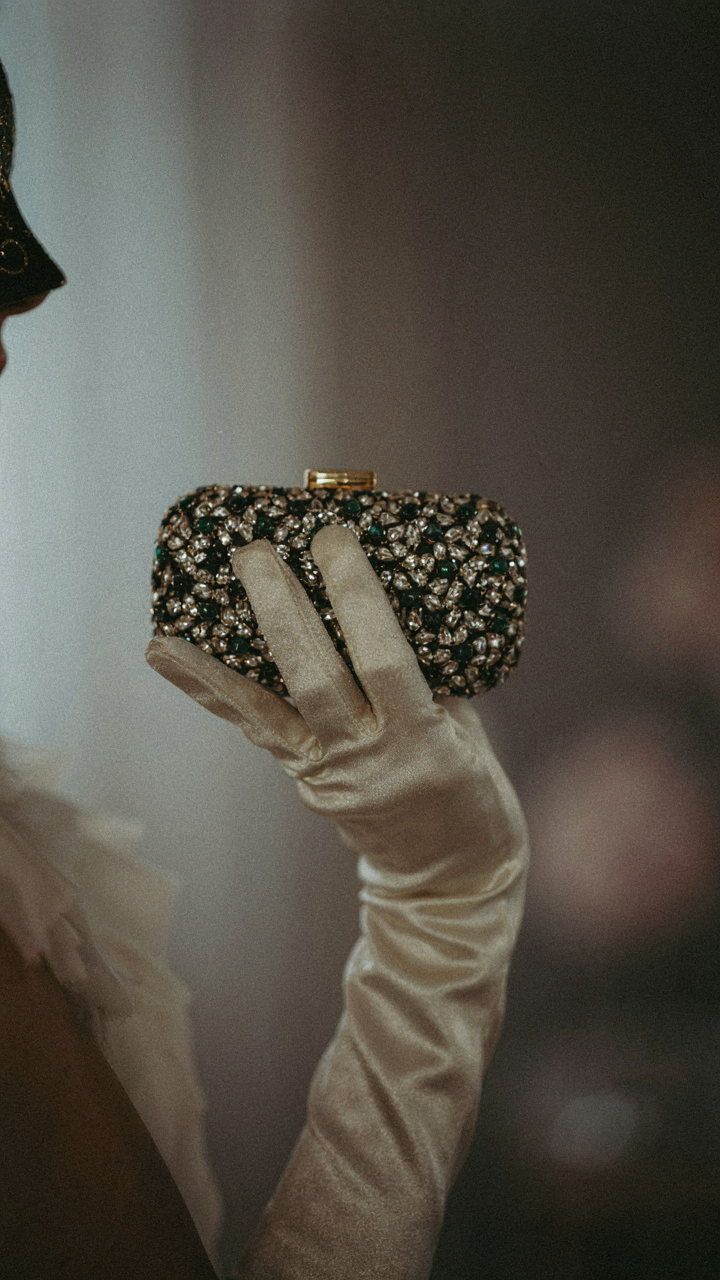 A masked woman's hand in a long glove, holding a jeweled clutch.