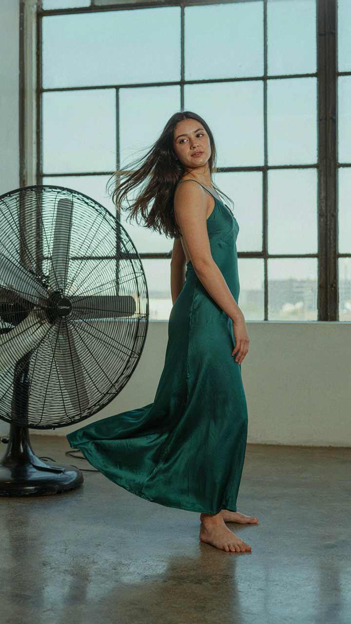 A woman in a satin dress stands near a fan, fabric gently moving.