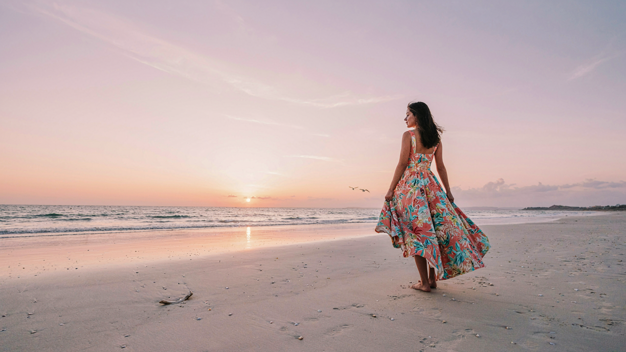 A woman in a flowing tropical wedding guest dress stands on a beach at sunset.