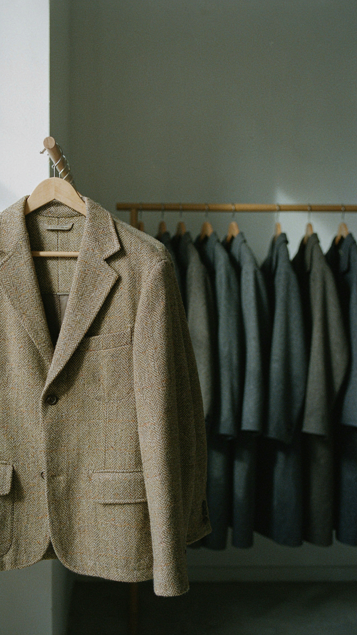 A tweed jacket stands out among many plain coats on a rack.