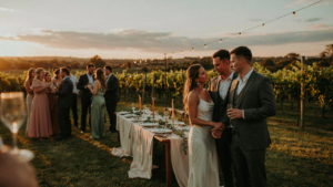A stylish couple smiling at a lively outdoor wedding welcome party as the sun sets.