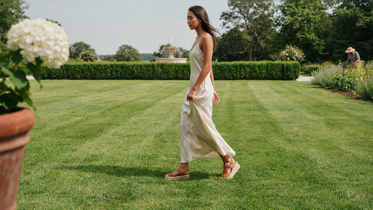 A woman in an elegant dress walks on a lawn, confidently wearing wedges for a garden wedding.