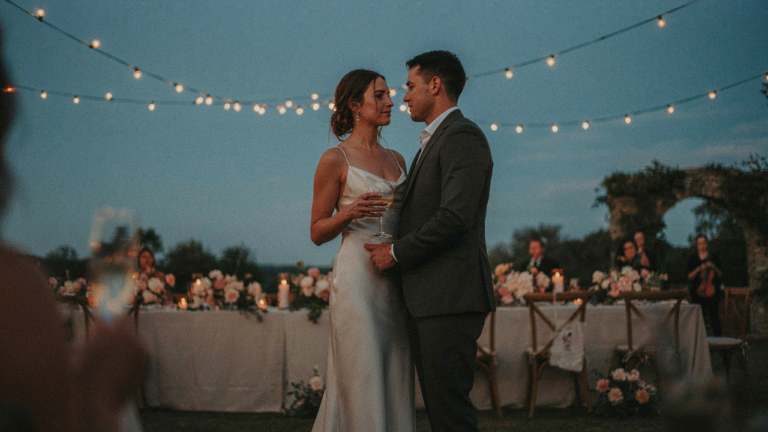 A couple in elegant cocktail attire stands together at an outdoor wedding reception.