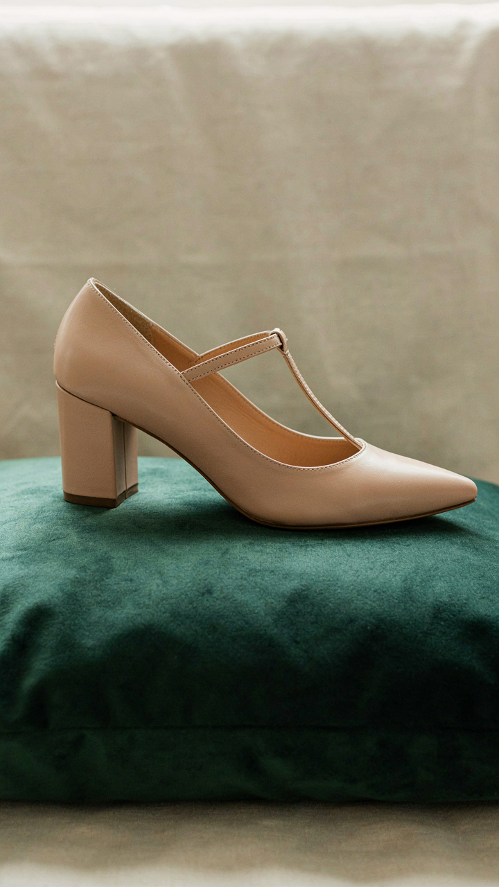 An elegant nude block heel with a metallic T-strap rests on velvet.