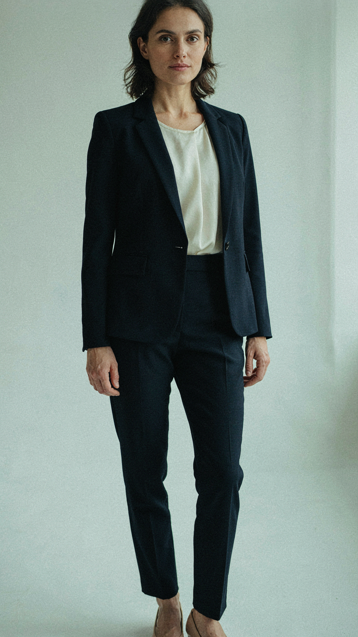 A woman wears a structured navy blazer, silk shell, tailored trousers, and flats.