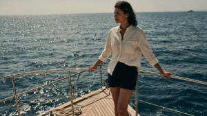 Woman in cream linen shirt and navy shorts stands confidently on a yacht deck.