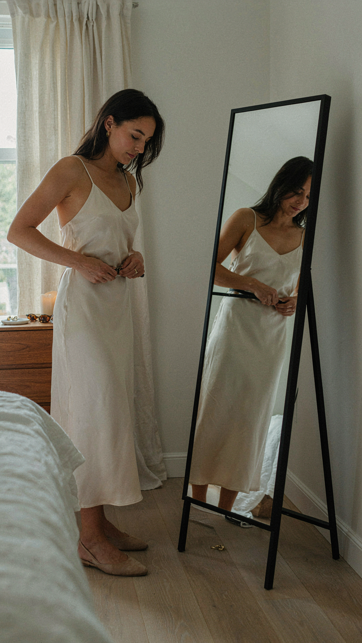 A woman looks in a mirror, adjusting a belt on her dress.