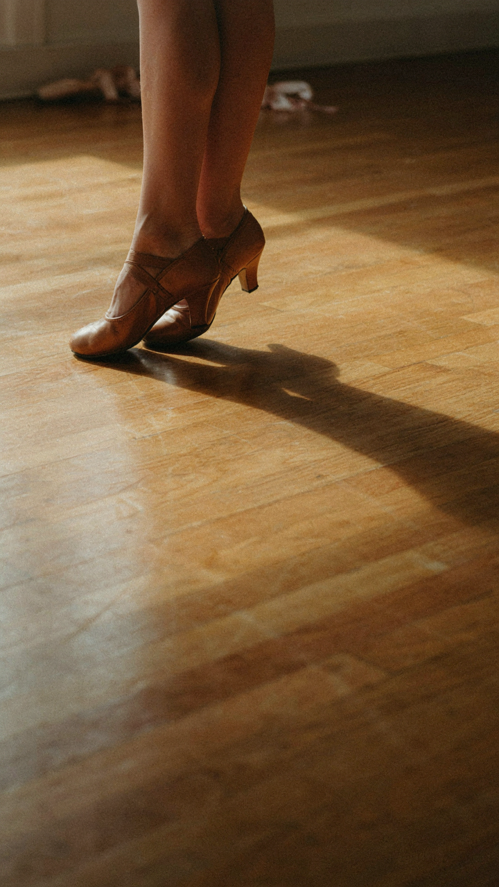 A person's feet in elegant dancing shoes cast a shadow on a polished dance floor.