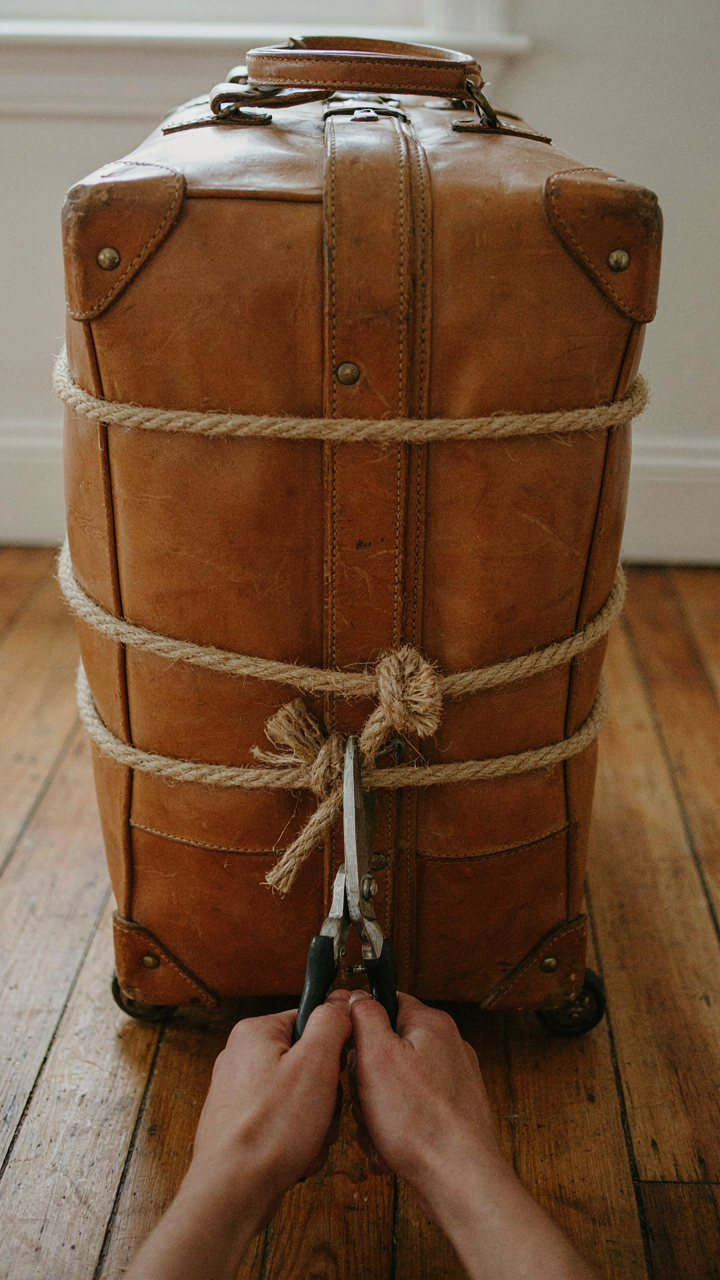 Hands cut a thick rope tied around a large, overstuffed suitcase with scissors.