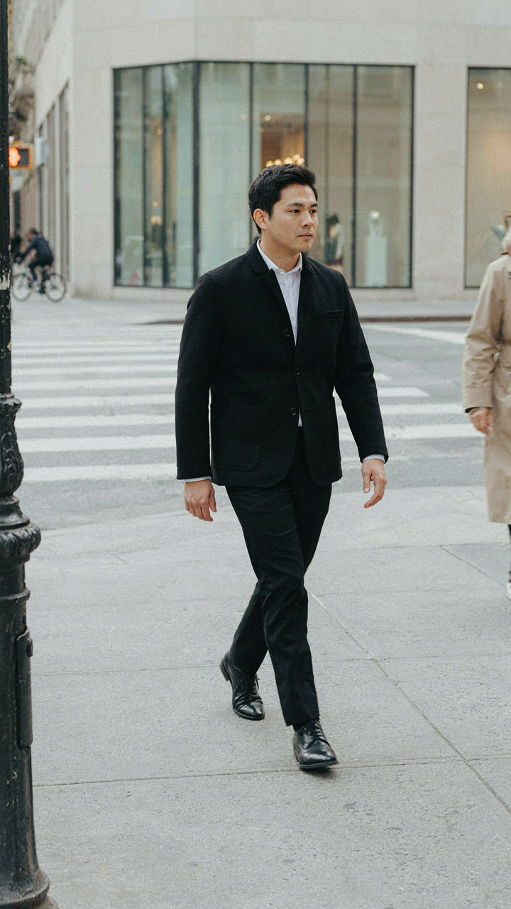 A person in a black blazer and tailored trousers walks confidently on a city street.